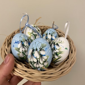 以下が含まれることがあります： 柳のバスケットには、手描きのイースターエッグが6個入っています。卵は、白と緑の花柄が薄い青または白の背景に描かれています。各卵には、吊り下げ用のリボンループが付いています。