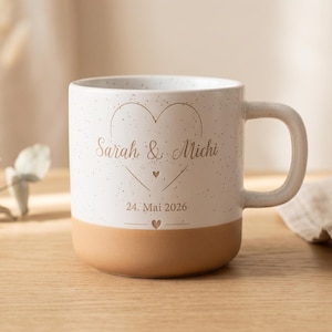 Könnte beinhalten: Eine weiße Keramiktasse mit gesprenkelter Oberfläche und einem beigefarbenen Boden. Auf der Vorderseite befindet sich ein brauner Aufdruck mit einem Herzumriss, den Namen Sarah & Michi, dem Datum 24. Mai 2026 und zwei kleinen Herzen. Mit Henkel.