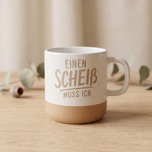Könnte beinhalten: Ein Keramikbecher mit gesprenkeltem weißem Oberteil und einem beigefarbenen Boden. Der Text EINEN SCHEIß MUSS ICH ist in Beigetönen auf der Vorderseite aufgedruckt. Der Becher hat einen geschwungenen Henkel an der rechten Seite und eine zylindrische Form.