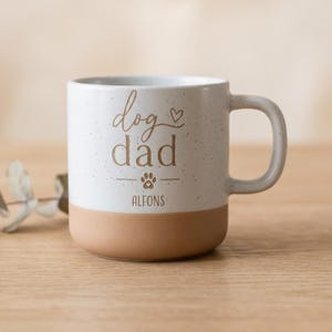 Könnte beinhalten: Ein weißer Keramikbecher mit gesprenkelter Oberfläche und beigem Boden. Auf der Vorderseite steht der Text "dog dad" in brauner Schrift, ergänzt durch ein kleines Herz, ein Pfotenabdruck-Symbol und den Namen "ALFONS" in braunen Buchstaben.