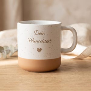 Könnte beinhalten: Eine Keramiktasse mit gesprenkeltem weißem Oberteil und einem einfarbigen beigen Boden. Auf der Vorderseite steht der Text "Dein Wunschtext" in brauner Schreibschrift über einem kleinen braunen Herzsymbol. Die Tasse hat einen geschwungenen Henkel.