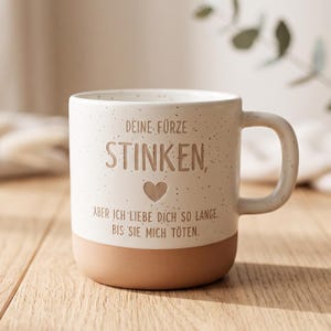 Könnte beinhalten: Eine cremefarbene Keramiktasse mit gesprenkelter Oberfläche und einem soliden terrakottafarbenen Boden. Auf der Vorderseite steht in brauner Schrift: DEINE FÜRZE STINKEN, ABER ICH LIEBE DICH SO LANGE. BIS SIE MICH TÖTEN. Ein kleines braunes Herz ist mittig.
