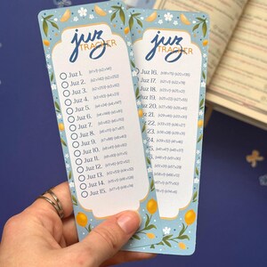 May include: Two blue "Juz Tracker" bookmarks with a floral and lemon design. Each bookmark has a list of Juz numbers with corresponding verse ranges. The text is in a stylized font. The bookmarks are held in a hand.