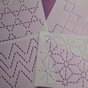 May include: Four white plastic stencils featuring geometric designs. Patterns include zigzags, diamonds, triangles, and starbursts, all with dotted lines. The stencils are arranged on a purple surface.