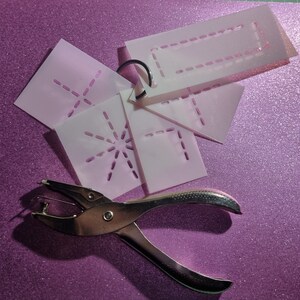 May include: A silver hole punch with a white plastic handle and four different stencils with dotted lines. The stencils are on a purple sparkly surface.