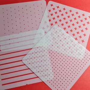 May include: Set of five white plastic stencils with different patterns. The patterns include polka dots, stripes, and a cross pattern. The stencils are on a red background.