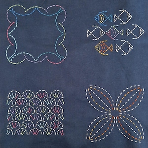 May include: A dark blue fabric with four different colorful embroidery designs. The designs include a square frame, a cluster of fish, a row of scallops, and a flower.