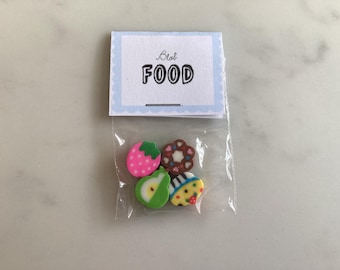 May include: A clear plastic bag containing four colorful eraser shapes: a strawberry, a pear, a flower, and a cupcake. The bag is sealed with a small paper tag that reads "Blah FOOD" in black letters, with a light blue scalloped border.