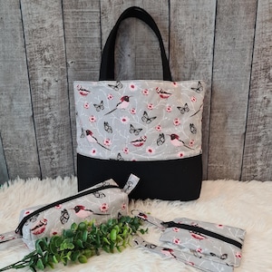 Könnte beinhalten: Eine Tragetasche und zwei passende Reißverschlusstaschen. Die Tragetasche hat einen schwarzen Boden und schwarze Griffe. Der obere Teil der Tasche und der Beutel haben einen grauen Hintergrund mit einem Muster aus rosa Vögeln, Schmetterlingen und Blumen. Die Beutel haben schwarze Reißverschlüsse.