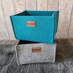 Könnte beinhalten: Zwei handgefertigte Filz-Aufbewahrungsboxen, eine türkis und eine graue, übereinander gestapelt. Jede Box hat ein Lederetikett mit der Aufschrift "HANDMADE". Die Boxen stehen auf einer strukturierten, grauen Oberfläche.