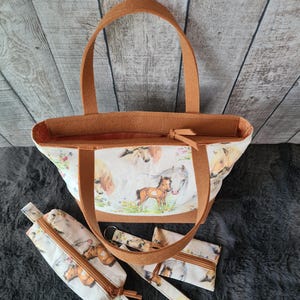 Könnte beinhalten: Eine Tragetasche, ein Federmäppchen, ein Schlüsselanhänger und eine kleine Geldbörse. Die Artikel haben einen weißen Hintergrund mit einem Pferdedruck. Die Tragetasche hat braune Griffe und Verzierungen. Das Federmäppchen, der Schlüsselanhänger und die Geldbörse haben braune Reißverschlüsse und Verzierungen.