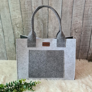 Könnte beinhalten: Graue Filz-Tragetasche mit einer rechteckigen Tasche auf der Vorderseite und zwei Griffen. Die Tasche hat ein kleines braunes Etikett mit der Aufschrift "HANDMADE". Die Tasche steht auf einer weißen, flauschigen Oberfläche.