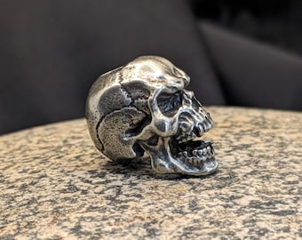 May include: A silver skull pendant with a detailed design. The skull is facing to the right and has an open mouth.