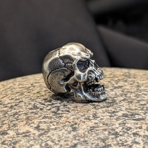 Könnte beinhalten: Ein silberner Totenkopf-Anhänger mit detailliertem Design. Der Schädel ist nach rechts gerichtet und hat einen offenen Mund.