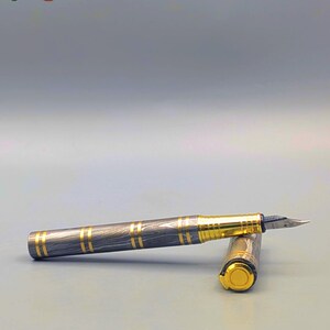 以下が含まれることがあります： 黒と金のデザインの万年筆。ペンは、金色のバンドと金色のペン先が付いた模様のバレルを持っています。キャップも金色です。画像には「Enigma」という文字があります。