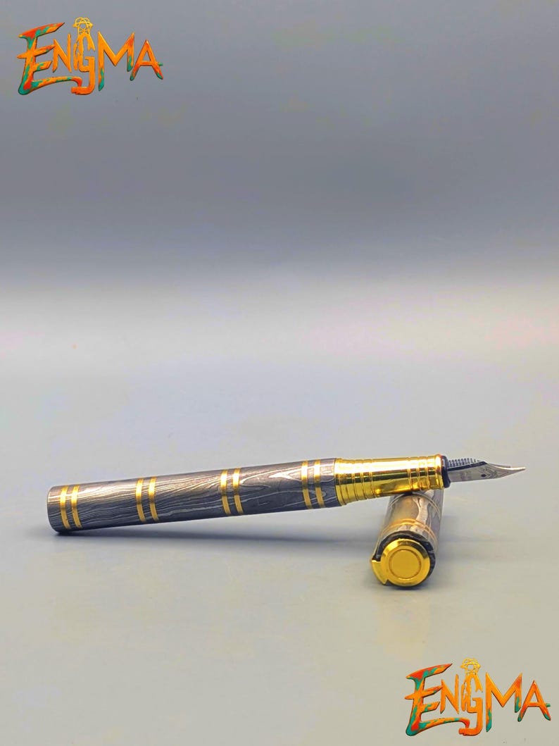 以下が含まれることがあります： 黒と金のデザインの万年筆。ペンは、金色のバンドと金色のペン先が付いた模様のバレルを持っています。キャップも金色です。画像には「Enigma」という文字があります。