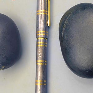 以下が含まれることがあります： ユニークな木目模様のシルバーとゴールドのペン。ペンにはゴールドのバンドとゴールドのクリップが付いています。「Enigma」の文字が画像左上と右下に表示されています。