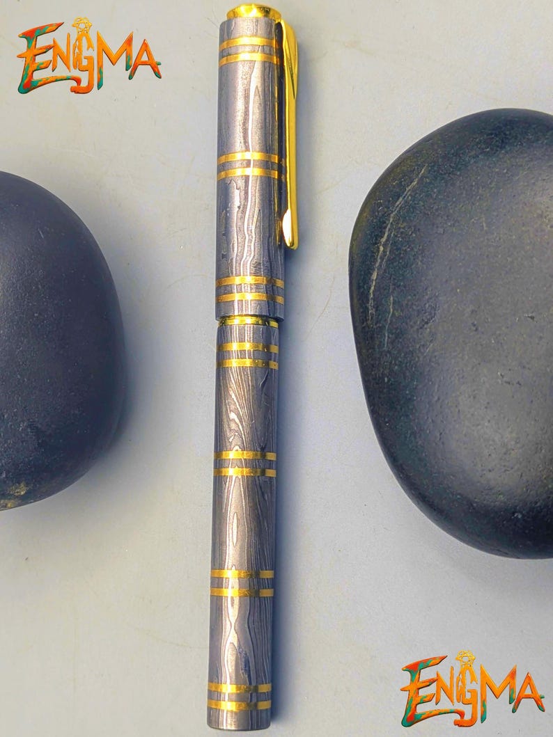 以下が含まれることがあります： ユニークな木目模様のシルバーとゴールドのペン。ペンにはゴールドのバンドとゴールドのクリップが付いています。「Enigma」の文字が画像左上と右下に表示されています。