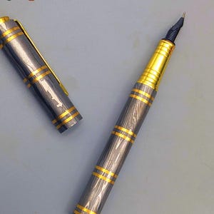 以下が含まれることがあります： グレーとゴールドのデザインの高級万年筆。ペンは、ゴールドのバンドとアクセントが付いたグレーのボディをしています。キャップは横にあります。ペンのペン先は黒と金です。「Enigma」という単語が左上と右下の隅にあります。