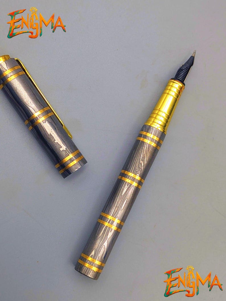 以下が含まれることがあります： グレーとゴールドのデザインの高級万年筆。ペンは、ゴールドのバンドとアクセントが付いたグレーのボディをしています。キャップは横にあります。ペンのペン先は黒と金です。「Enigma」という単語が左上と右下の隅にあります。