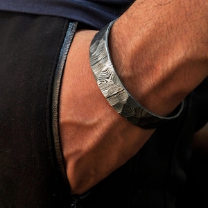 Könnte beinhalten: Ein silberfarbenes Metallarmband mit einem einzigartigen Muster. Das Armband hat eine strukturierte Oberfläche mit einem wellenartigen Muster. Das Armband wird am Handgelenk einer Person getragen, mit einem dunkelblauen Hemd und einer schwarzen Hose im Hintergrund.