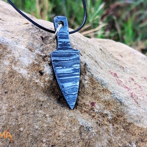 Könnte beinhalten: Ein silberner Anhänger in Form einer Speerspitze mit einem schwarzen Lederband. Der Anhänger hat ein Muster aus wellenförmigen Linien, was ihm ein einzigartiges Aussehen verleiht.