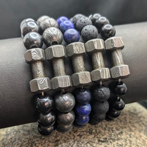 May include: A stack of bracelets featuring black, blue, and gray beads. The bracelets are made of various materials, including lava rock, metal, and gemstone beads.