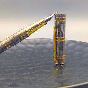 以下が含まれることがあります： 金色のペン先とアクセントが付いた万年筆。ペンの本体は濃いグレーとゴールドの模様です。ペンのキャップが開いており、ペン先が見えています。「Enigma」の文字が左上と右下に表示されています。