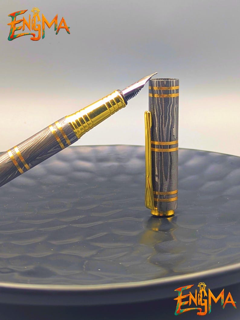 以下が含まれることがあります： 金色のペン先とアクセントが付いた万年筆。ペンの本体は濃いグレーとゴールドの模様です。ペンのキャップが開いており、ペン先が見えています。「Enigma」の文字が左上と右下に表示されています。