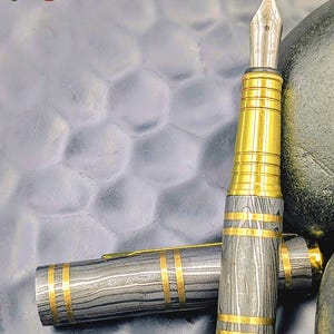 以下が含まれることがあります： 銀色のペン先と金色のアクセントが特徴の高級万年筆。ペンのボディは、金色のバンドで補完された、グレーと黒の独特の縞模様が特徴です。「Enigma」の文字が左上と右下に表示されています。