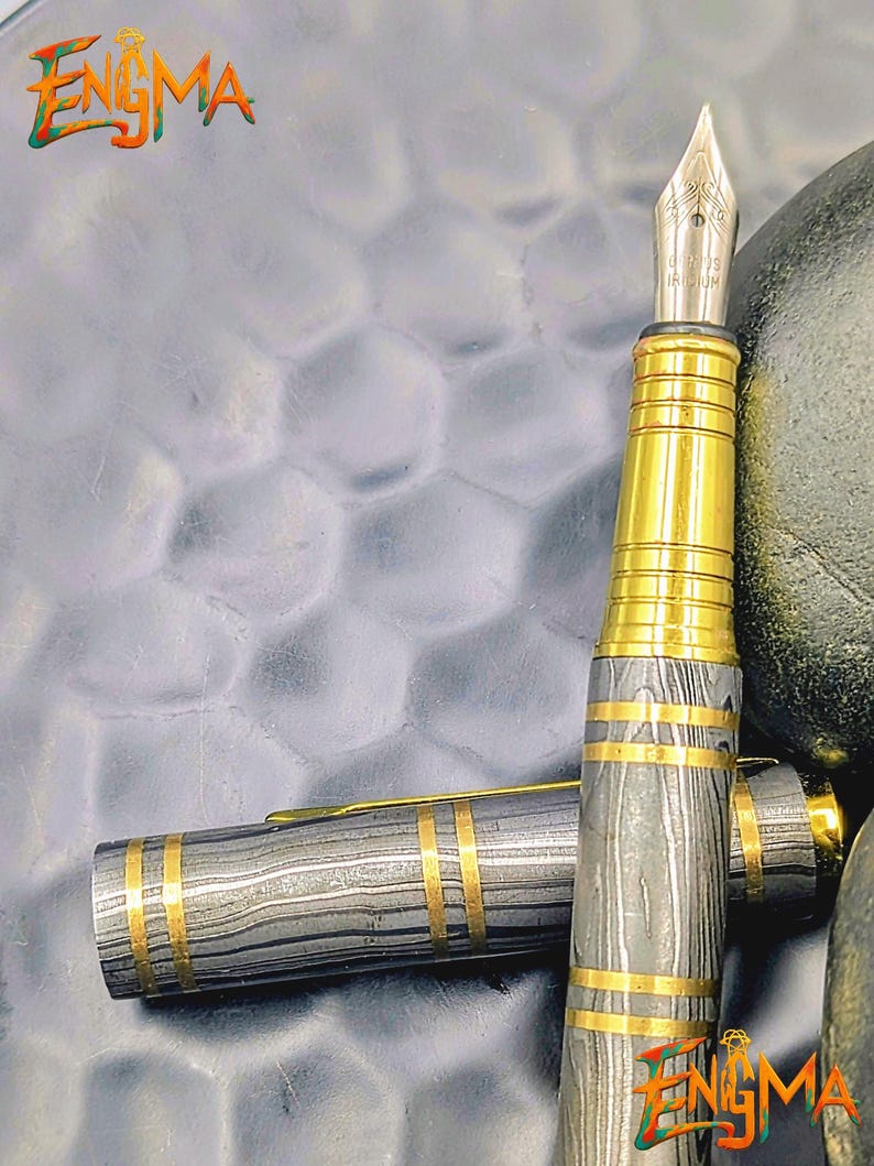 以下が含まれることがあります： 銀色のペン先と金色のアクセントが特徴の高級万年筆。ペンのボディは、金色のバンドで補完された、グレーと黒の独特の縞模様が特徴です。「Enigma」の文字が左上と右下に表示されています。