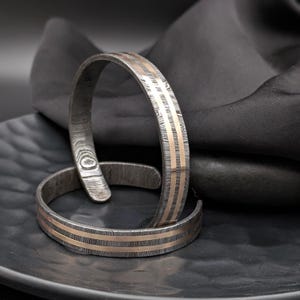 May include: Two metal cuff bracelets. The bracelets have a textured, patterned appearance with two parallel gold-colored stripes. The bracelets are displayed on a dark, textured surface.