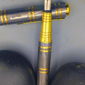 以下が含まれることがあります： 金色のペン先とアクセントが付いた高級万年筆。ペンは、模様のあるグレーとゴールドのバレルとキャップが特徴です。「Enigma」の文字が左上と右下に表示されています。