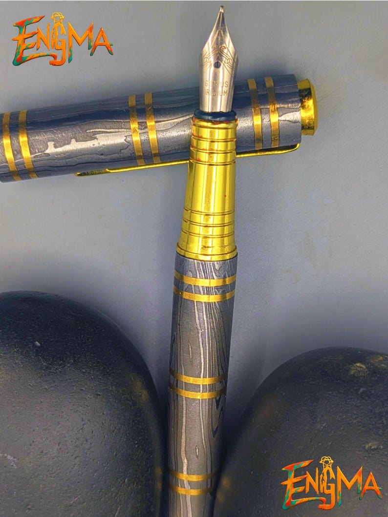 以下が含まれることがあります： 金色のペン先とアクセントが付いた高級万年筆。ペンは、模様のあるグレーとゴールドのバレルとキャップが特徴です。「Enigma」の文字が左上と右下に表示されています。
