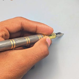 以下が含まれることがあります： グレーとゴールドの模様が入った万年筆が、水色の背景に持たれています。ペン先は金で、金色のバンドが付いています。「Enigma」の文字がカラフルで様式化されたフォントで表示されています。