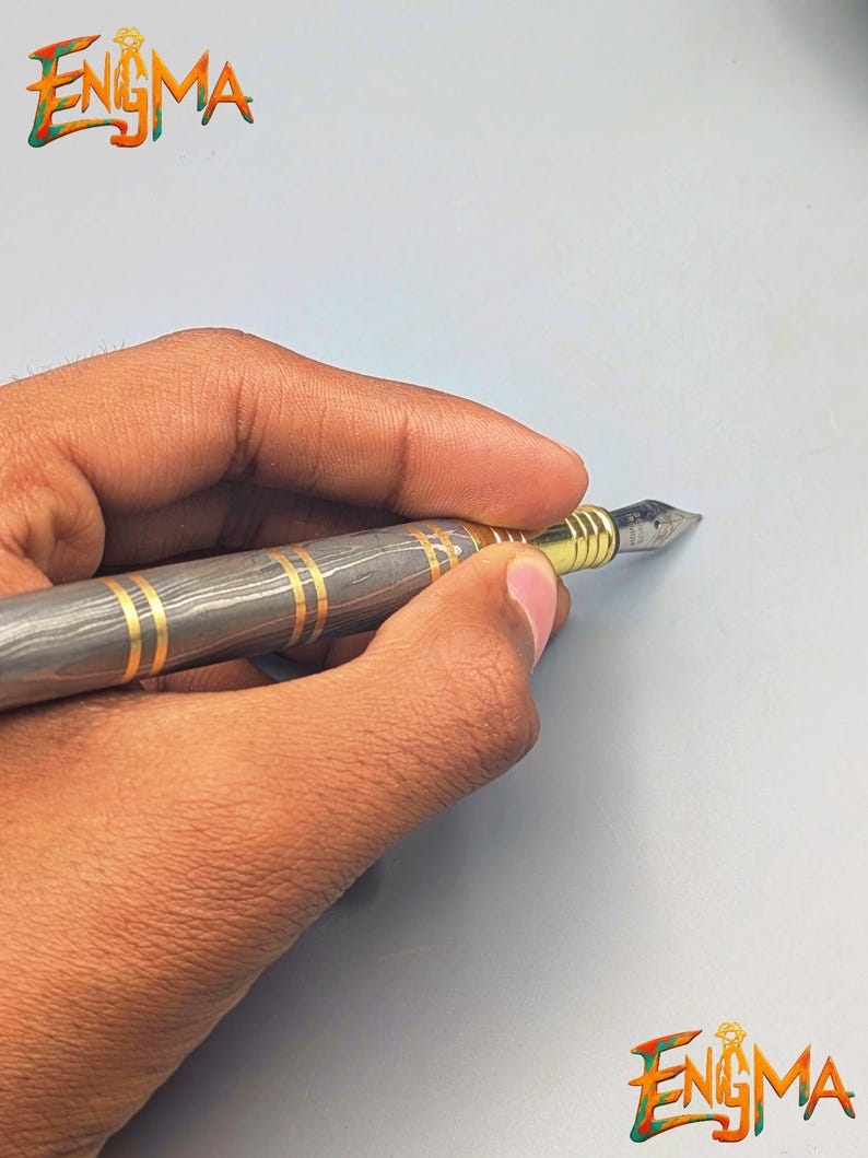 以下が含まれることがあります： グレーとゴールドの模様が入った万年筆が、水色の背景に持たれています。ペン先は金で、金色のバンドが付いています。「Enigma」の文字がカラフルで様式化されたフォントで表示されています。