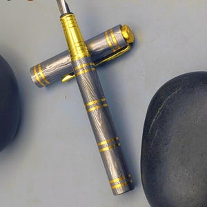 以下が含まれることがあります： 銀と金のデザインの高級万年筆。 ペンは、金色のアクセントと金色のペン先が付いた銀色のボディが特徴です。 キャップは外れており、ペン先が見えています。 「Enigma」の文字が確認できます。