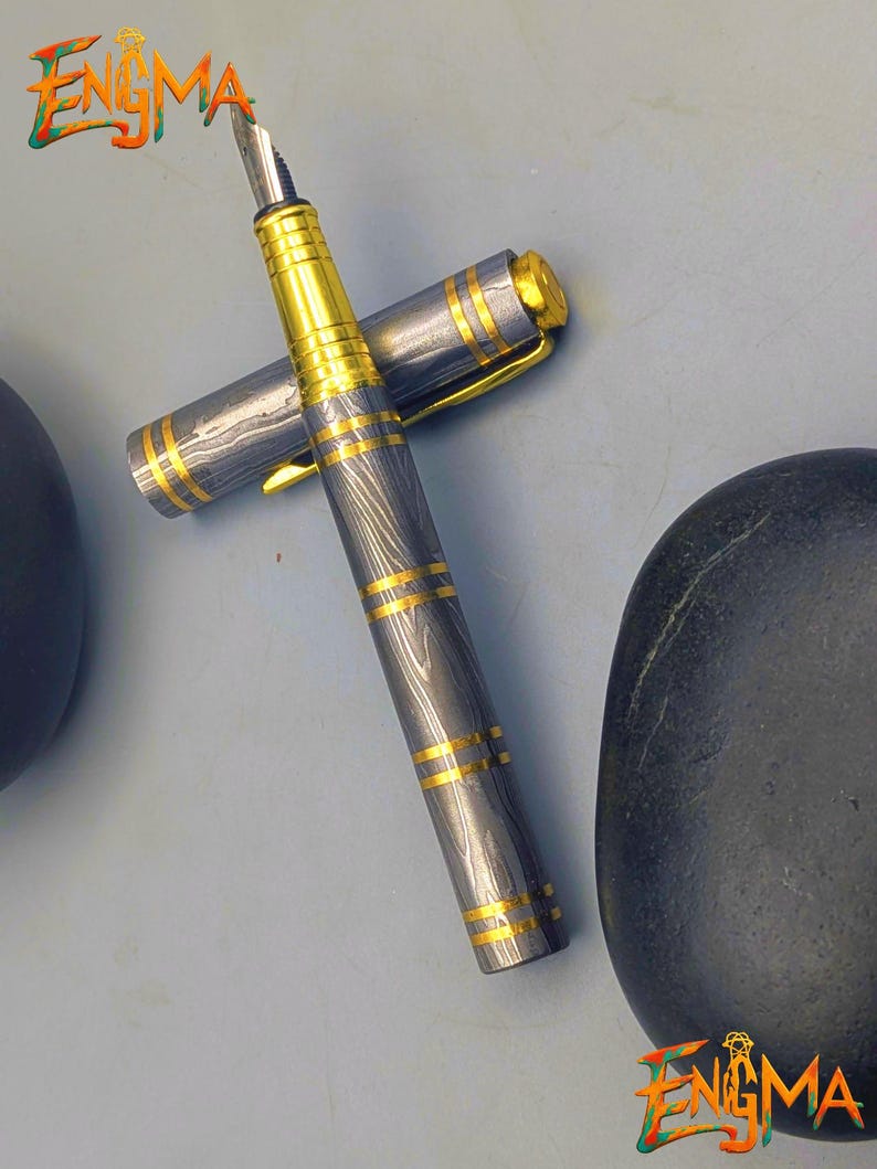 以下が含まれることがあります： 銀と金のデザインの高級万年筆。 ペンは、金色のアクセントと金色のペン先が付いた銀色のボディが特徴です。 キャップは外れており、ペン先が見えています。 「Enigma」の文字が確認できます。