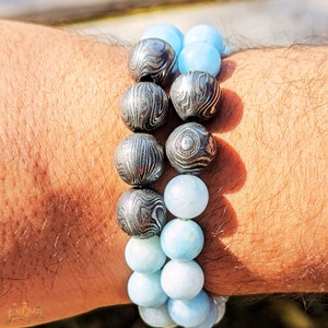 May include: Two bracelets stacked on a wrist. The top bracelet is made of dark gray patterned beads. The bottom bracelet is made of light blue beads.