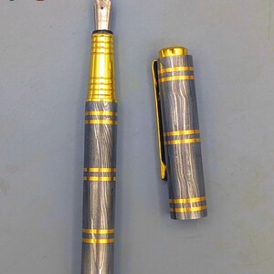 以下が含まれることがあります： 銀色のペン先とグレーとゴールドのボディの万年筆。ペンのボディには、金色のバンドが付いた木目模様があります。キャップは外れており、ペンのペン先が見えています。「Enigma」の文字が左上と右下にあります。