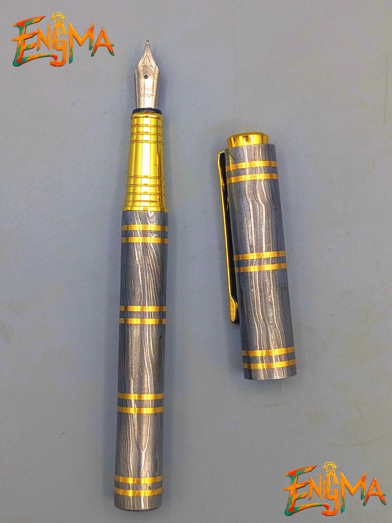 以下が含まれることがあります： 銀色のペン先とグレーとゴールドのボディの万年筆。ペンのボディには、金色のバンドが付いた木目模様があります。キャップは外れており、ペンのペン先が見えています。「Enigma」の文字が左上と右下にあります。