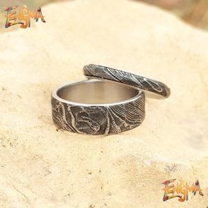 May include: Two textured metal rings. The larger ring has a wide band with a swirling pattern, while the smaller ring has a thinner band with a linear design. The rings are displayed on a light brown surface. The word "Enigma" is in the image.