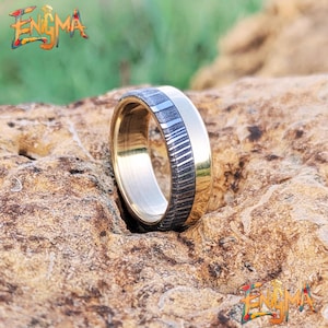 Könnte beinhalten: Ein goldfarbener Ring mit einem kontrastierenden Band aus Dunkelgrau und Silber, platziert auf einer strukturierten Steinoberfläche. Der Ring zeichnet sich durch ein einzigartiges Design mit sichtbarer Handwerkskunst aus. Das Wort "Enigma" ist in einer farbenfrohen, stilisierten Schrift dargestellt.