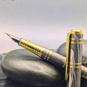 以下が含まれることがあります： 金色のディテールと独特の黒とグレーの模様が特徴の高級万年筆。画像左上と右下に「Enigma」の文字が表示されています。ペンのキャップは外されています。