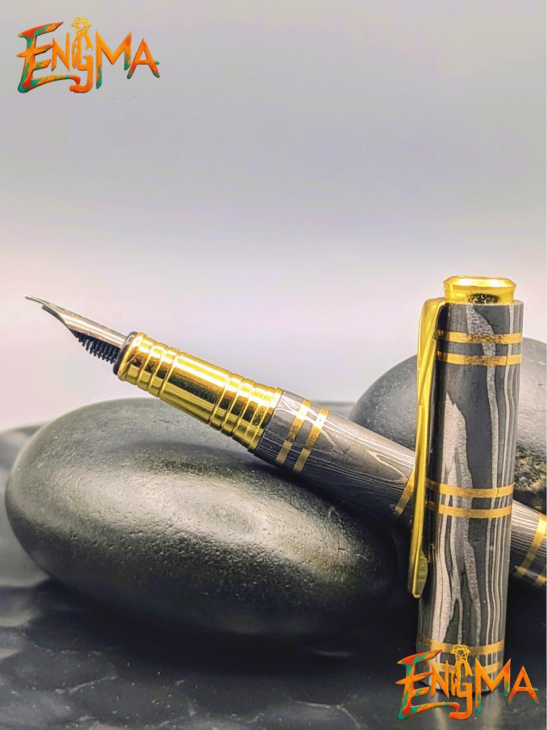以下が含まれることがあります： 金色のディテールと独特の黒とグレーの模様が特徴の高級万年筆。画像左上と右下に「Enigma」の文字が表示されています。ペンのキャップは外されています。