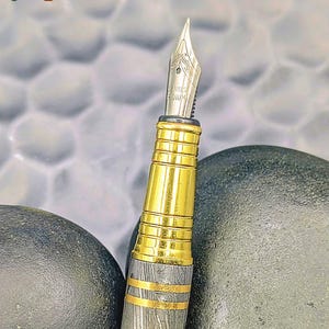 以下が含まれることがあります： 金色のボディと銀色のペン先を備えた高級万年筆のクローズアップ。ペンには複雑なディテールが施されており、暗い背景に配置されています。「Enigma」の文字が左上と右下に表示されています。