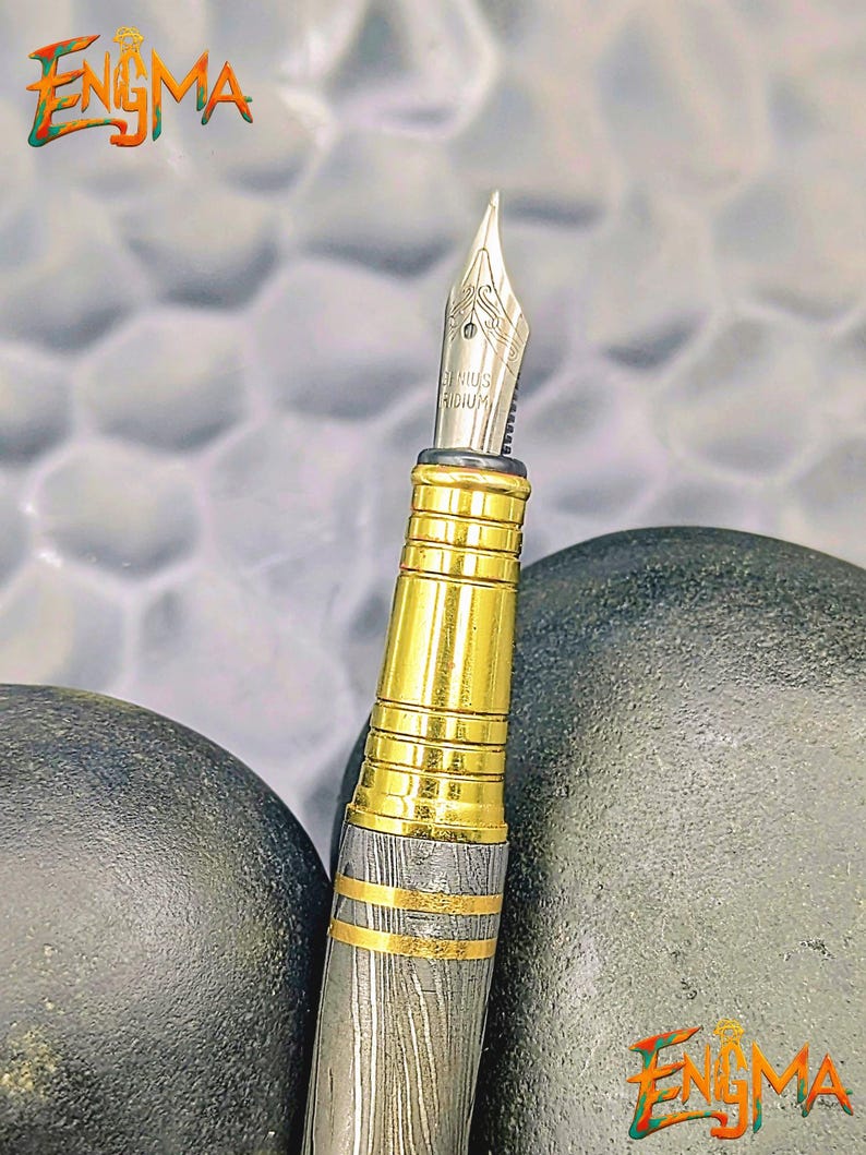 以下が含まれることがあります： 金色のボディと銀色のペン先を備えた高級万年筆のクローズアップ。ペンには複雑なディテールが施されており、暗い背景に配置されています。「Enigma」の文字が左上と右下に表示されています。