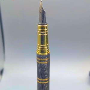 以下が含まれることがあります： 黒と金のペン先を備えた万年筆のクローズアップ。ペンの本体は、金色のバンドが付いた濃い灰色の木目模様が特徴です。「Enigma」の文字がカラフルで様式化されたフォントで表示されています。