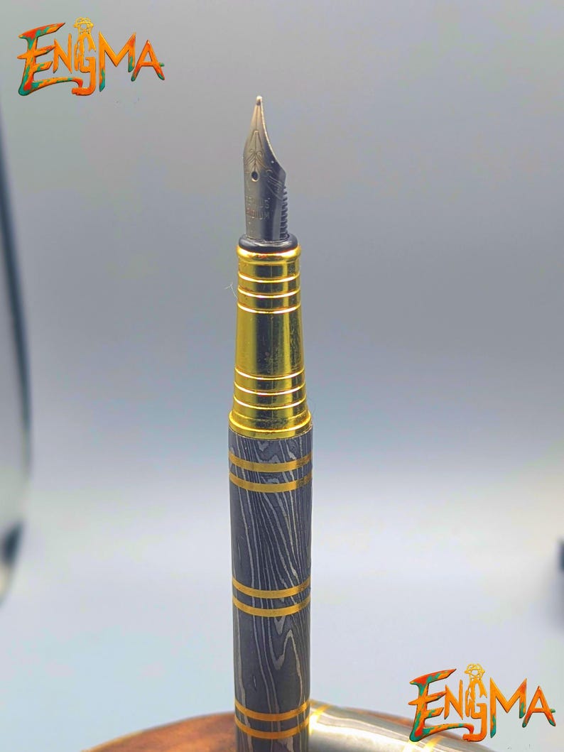 以下が含まれることがあります： 黒と金のペン先を備えた万年筆のクローズアップ。ペンの本体は、金色のバンドが付いた濃い灰色の木目模様が特徴です。「Enigma」の文字がカラフルで様式化されたフォントで表示されています。