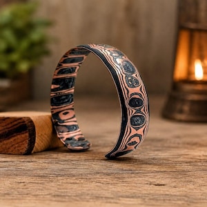 May include: A cuff bracelet featuring a layered mokume gane pattern with alternating bands of dark charcoal grey and metallic copper. The metal surface displays a swirling, organic aesthetic with a smooth, polished finish and an open-ended design.
